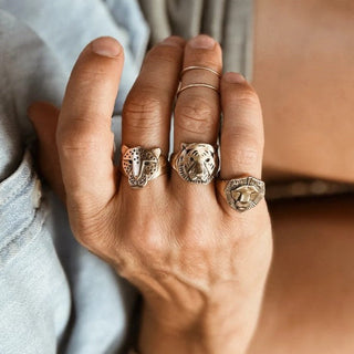 A close-up of a hand wearing three silver rings with animal head designs, including a lion and a tiger, along with a thin gold band and a silver bracelet.