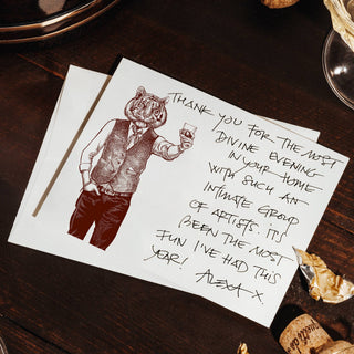 A square white greeting card with a hand-drawn illustration of a cat in a vest and a handwritten thank-you message, styled on a dark wooden table with wine glasses, wine bottles, and gold foil.