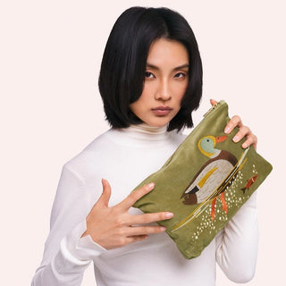 A woman with short black hair wearing a white turtleneck holds a sage green velvet pouch decorated with a detailed embroidered duck design and small floral accents, set against a solid pale pink background.