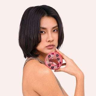 A woman with short dark hair poses in a brown and beige checkered sleeveless dress, holding a pink frosted donut with dark berry sprinkles against her shoulder, set against a solid pale pink background.