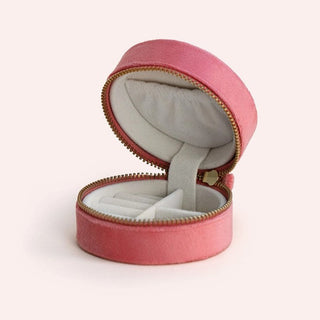 An open round pink velvet jewelry box with a white velvet interior and gold-tone zipper, set against a solid pale pink background.
