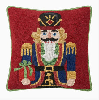 A square red needlepoint pillow featuring a detailed, colorful embroidered nutcracker soldier design with a green border on a white background.