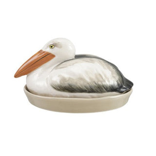 A ceramic pelican figurine sitting in a shallow oval dish, with a white head and body, gray wings, and a tan-orange beak, shown on a plain white background.