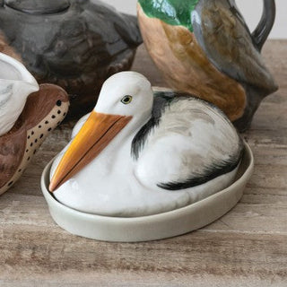 A ceramic pelican figurine in a shallow oval dish, displayed on a rustic wooden surface with other decorative ceramic items blurred in the background.