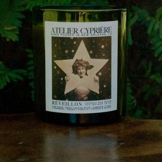 A dark glass candle jar with a white label reading 'ATELIER CYPRIÈRE' and a vintage-style star illustration, set on a wooden surface with green foliage in the background.