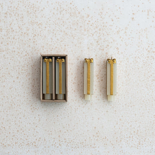 Three gold-tone and off-white cylindrical earrings displayed next to their matching cardboard packaging on a speckled white background.