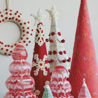 A collection of decorative Christmas tree ornaments and tree-shaped decor pieces arranged on a tabletop against a light neutral wall. The set includes red, white, and pink felt cone trees with embroidered details, translucent red and pink tiered tree ornaments, a small mint green tree ornament, and a white wreath with red polka dots hanging in the background.
