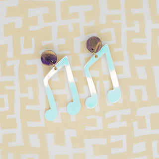 A pair of dangling earrings shaped like AQUA BLUE musical notes with white stems and small metallic gold-toned studs, placed on a pale yellow background with a white maze pattern.
