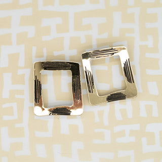 A pair of gold-toned acrylic square stud earrings with a textured, ribbed surface, displayed on a cream and white geometric patterned background.