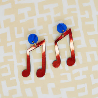 A pair of novelty earrings shaped like red musical notes with blue circular studs, displayed on a light beige background with a white maze pattern.