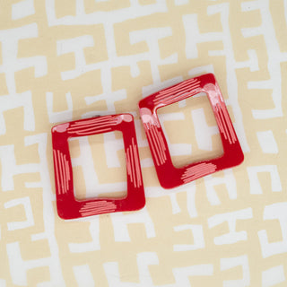 A pair of red square frame acrylic earrings with white line-art patterns, displayed on a light beige background with a white geometric maze pattern.