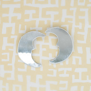 Two silver crescent-shaped acrylic earrings arranged to form a circular shape, placed on a beige background with a white maze-like pattern.