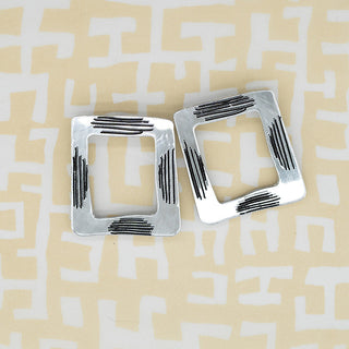 A pair of silver square earrings with black textured stripes, displayed on a beige and white patterned background.