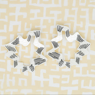 Two white squiggle shape earrings with black striped patterns, arranged on a pale yellow background with a white maze-like print.