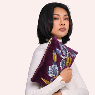 A woman with short black hair wearing a white long-sleeve top holds a purple velvet pouch bag decorated with a blue, white, and gold floral pattern against a light beige background.