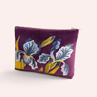 A purple velvet cosmetic pouch with embroidered irises in white, blue, and yellow, featuring a gold zipper closure, set against a solid pale pink background.