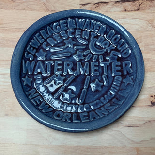 A round ceramic watermeter mini dish with embossed text and decorative patterns, primarily in a gray color, placed on a wooden surface.